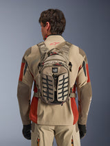 Alpinestars AT-15 Back Pack, Aluminum Walnut, technical adventure motorcycle backpack worn by a rider in a beige and red jacket, featuring tire-tread pattern, TERRA logo, red zipper pulls, and adjustable straps