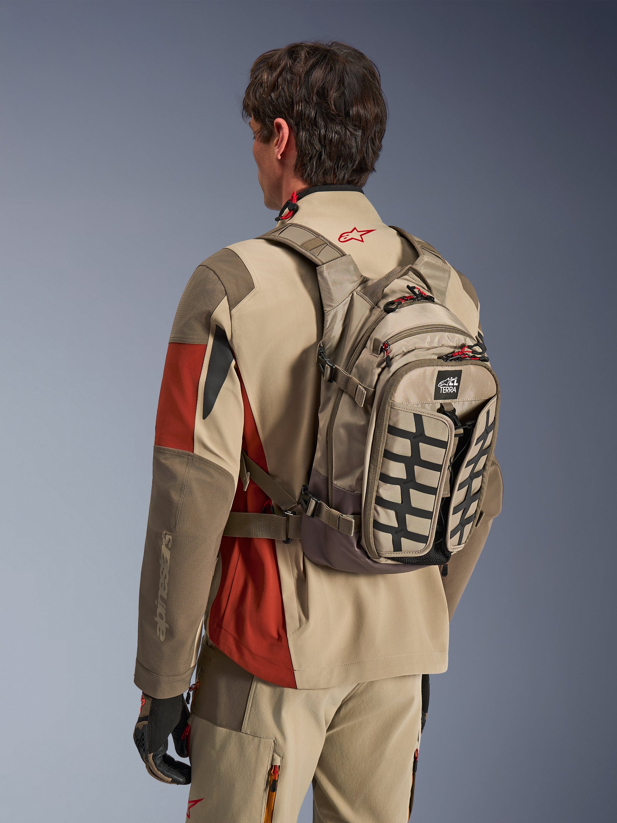 Alpinestars AT-15 Back Pack in Aluminum Walnut, technical motorcycle backpack worn by a rider in matching adventure gear, featuring tan and brown textile construction with black structural accents and red zipper pulls, designed for off-road and adventure riding.