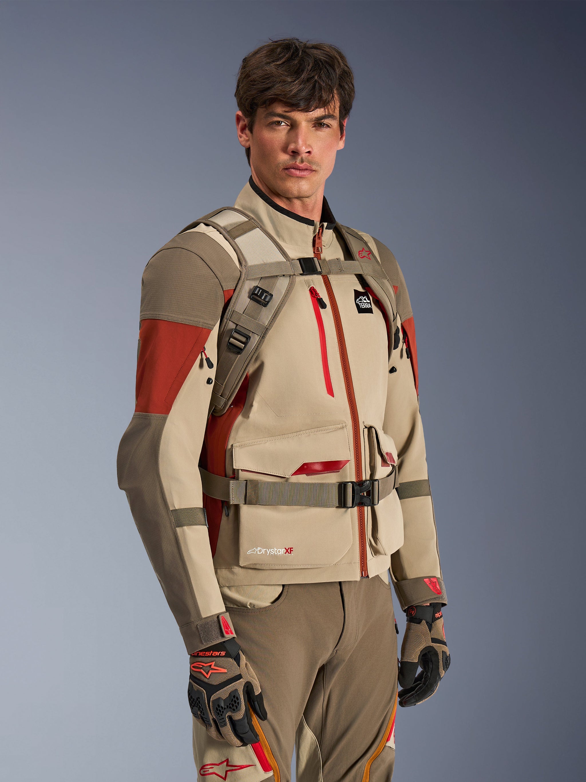 Alpinestars AT-15 Back Pack in Aluminum Walnut, worn by a rider over a matching adventure motorcycle jacket, showing the front harness with magnetic chest lock and adjustable straps, tan and red technical gear for adventure riding