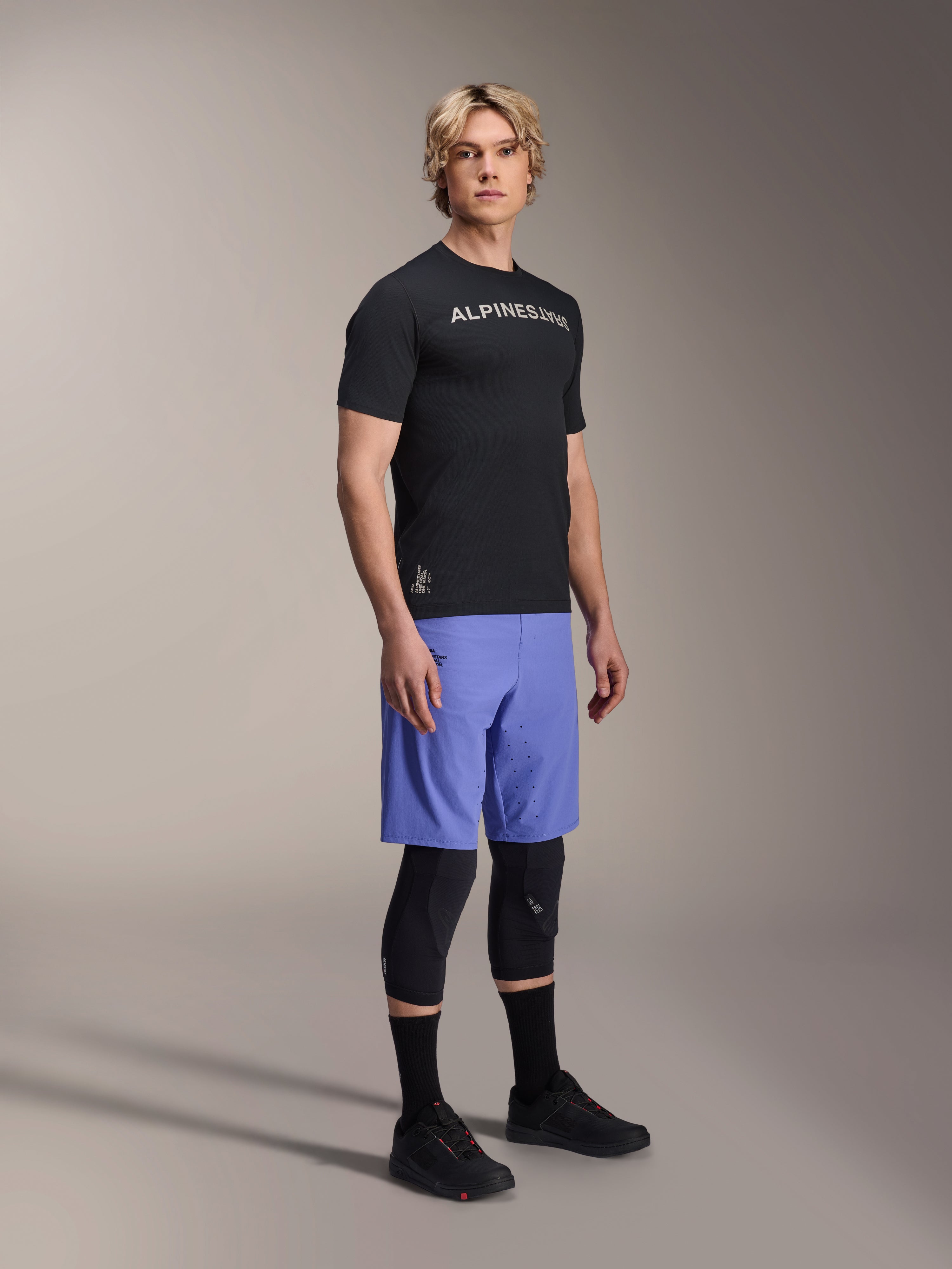 Male model wearing Alpinestars A-Aria Switch Short Sleeve Jersey in Black with white chest branding, paired with light blue mountain bike shorts, black knee guards, and black cycling shoes, studio full-body shot for performance MTB gear