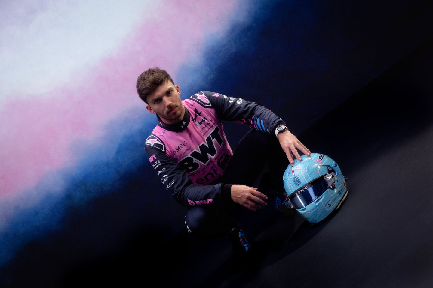 Pierre Gasly in an Alpinestars auto racing suit holding a helmet with colorful smoke effect on a black, pink and blue background. 