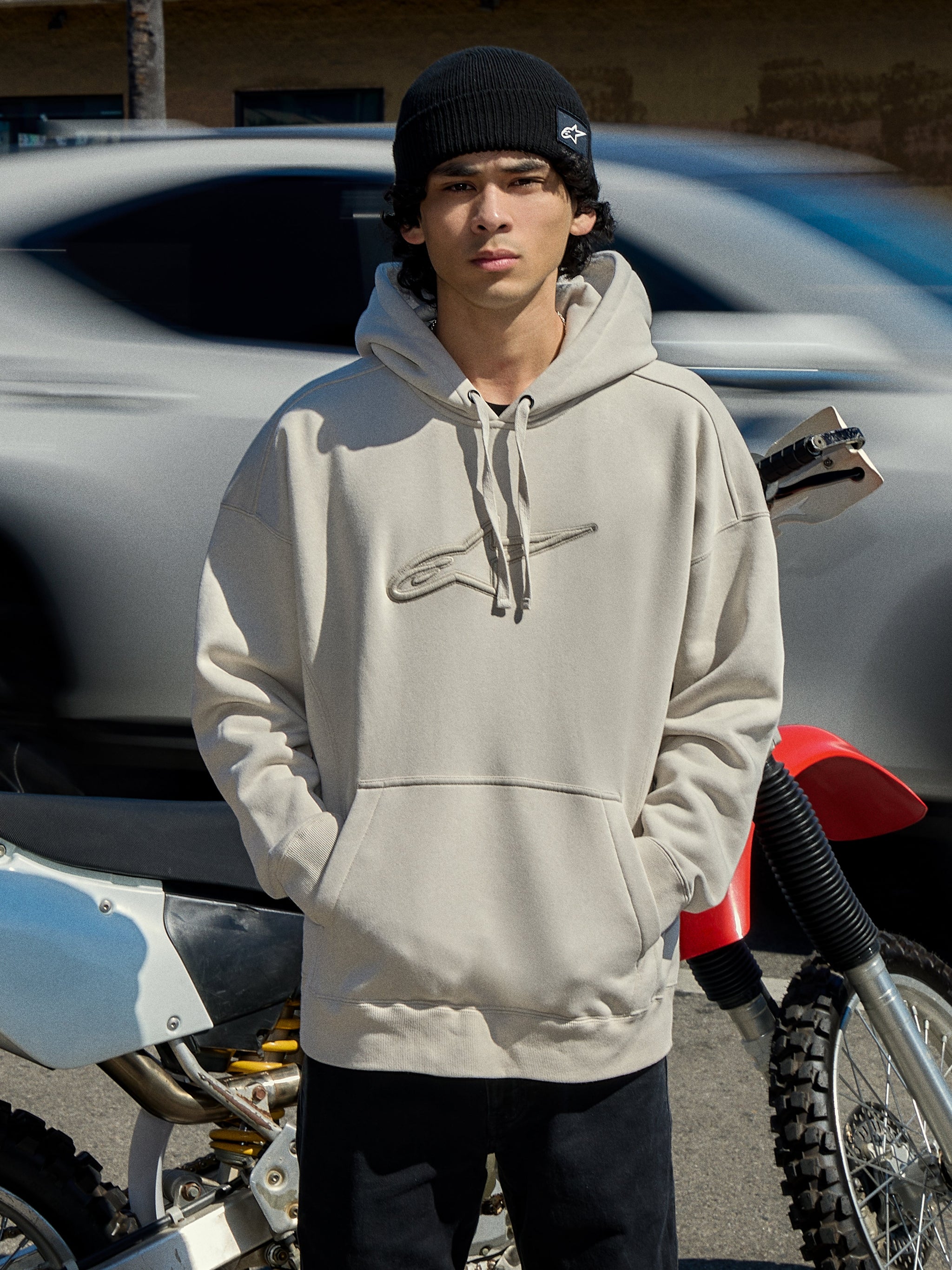 Alpinestars Emergent Oversized Hoodie in Off White, oversized pullover fleece with 3D embroidered Astars logo, kangaroo pocket, and drawstring hood, worn by a man with a black beanie next to a motorcycle, casual lifestyle motorsport streetwear