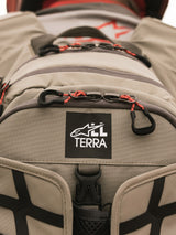 Alpinestars AT-15 Back Pack, Aluminum Walnut, close-up view of All Terra logo patch on tan technical textile, featuring orange corded zipper pulls and rugged construction details