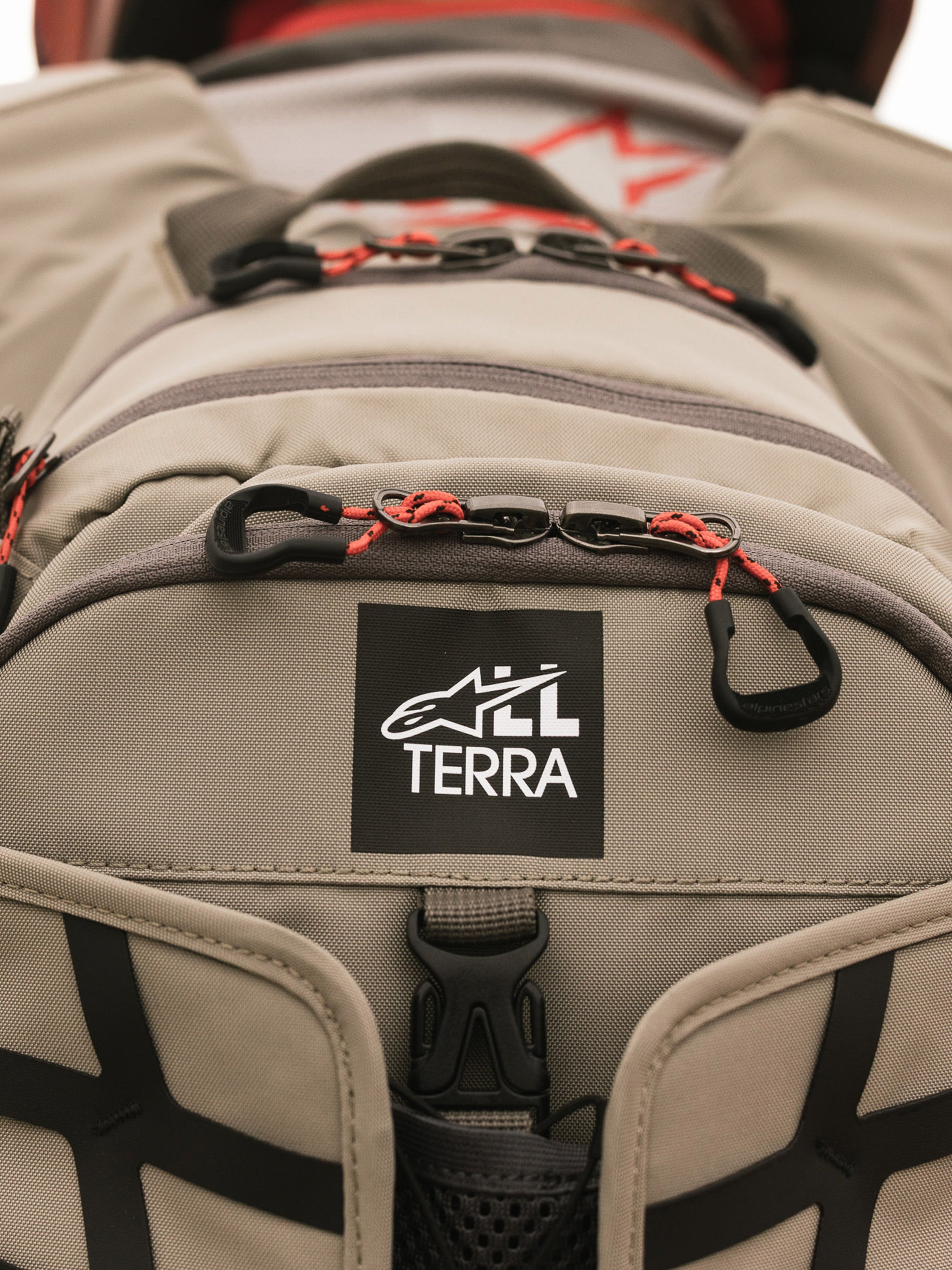 Alpinestars AT-15 Back Pack, Aluminum Walnut, close-up view of All Terra logo patch on tan technical textile, featuring orange corded zipper pulls and rugged construction details