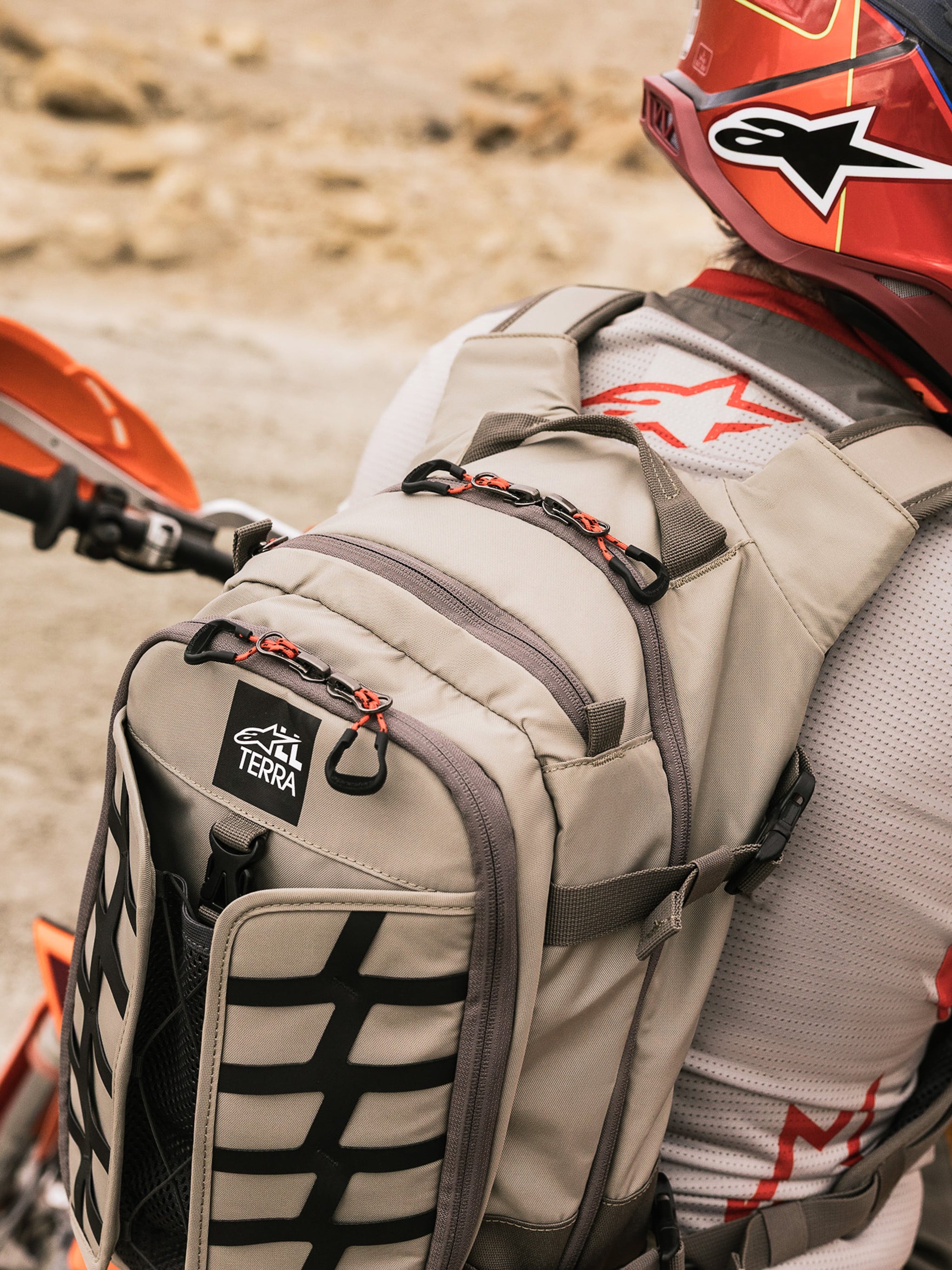 Alpinestars AT-15 Back Pack, Adventure Motorcycle Backpack, Aluminum Walnut and Black, worn by a rider in a desert setting, featuring red zipper pulls, TERRA logo patch, and black external webbing, paired with a matching red helmet and white Alpinestars jersey