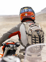 Alpinestars AT-15 Back Pack in Aluminum Walnut, worn by an adventure motorcycle rider in a desert landscape. Features rugged beige textile construction with black webbing, orange zipper accents, and 'All Terra' branding. Paired with a red and orange helmet and grey riding gear for off-road touring.