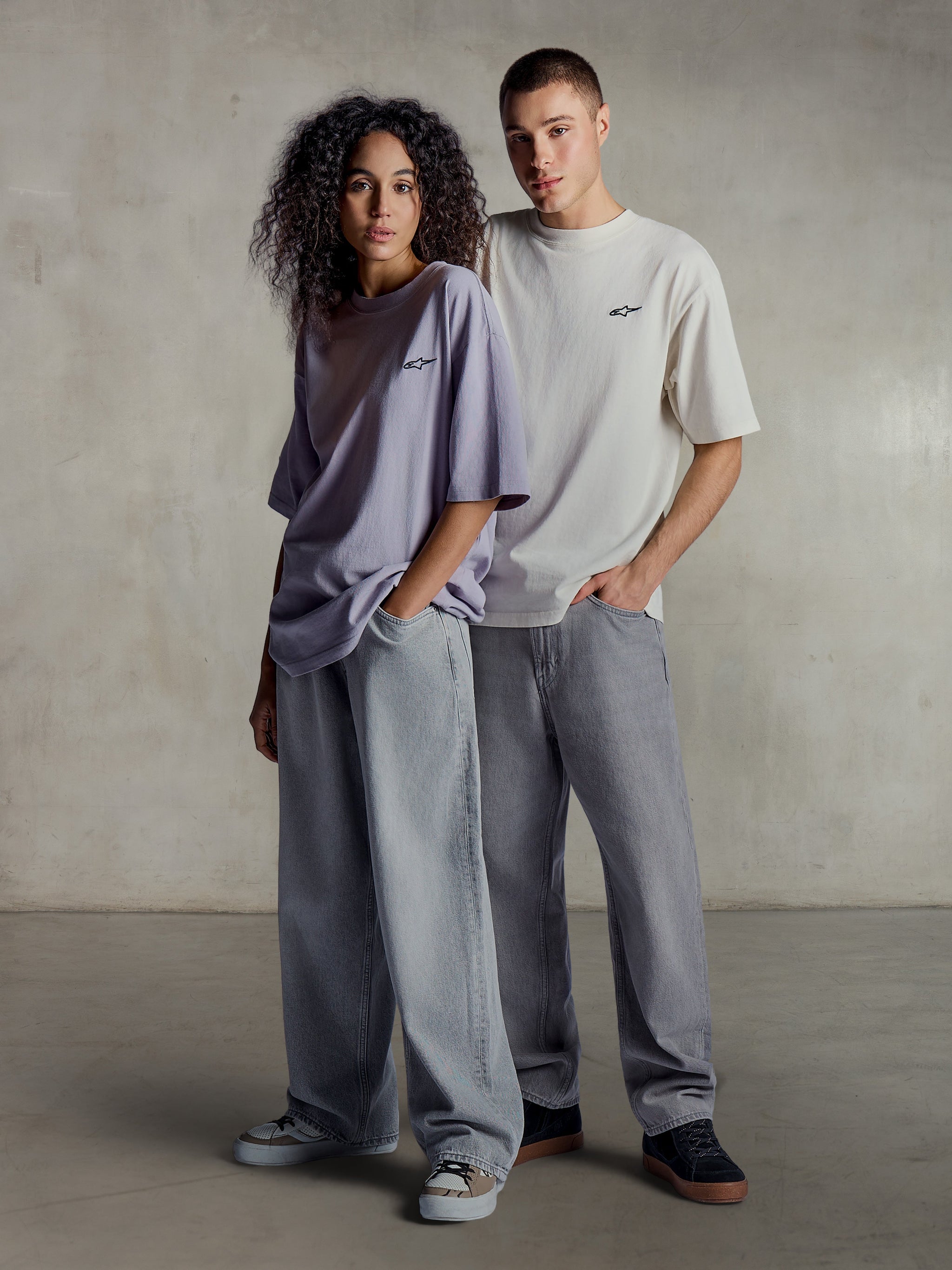 Alpinestars Essentials Ageless SS Tee, lifestyle view of two models wearing short sleeve tees in light purple and silver, relaxed fit organic cotton with a subtle chest logo, paired with loose jeans for casual streetwear style.