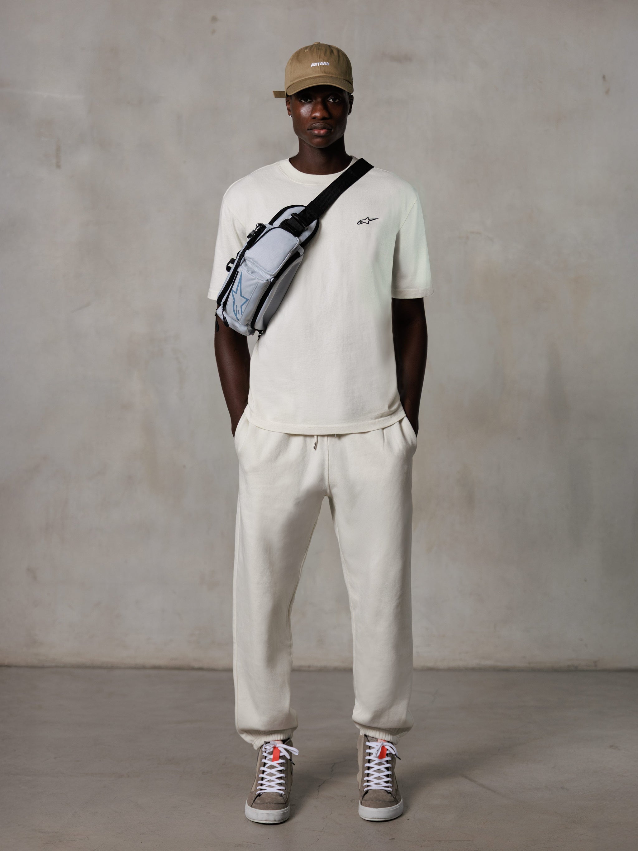 Alpinestars Essentials Ageless SS Tee, Short Sleeve T-Shirt, Off White, featuring a small black logo on the chest, worn with matching joggers, tan baseball cap, and a grey crossbody bag, casual lifestyle streetwear