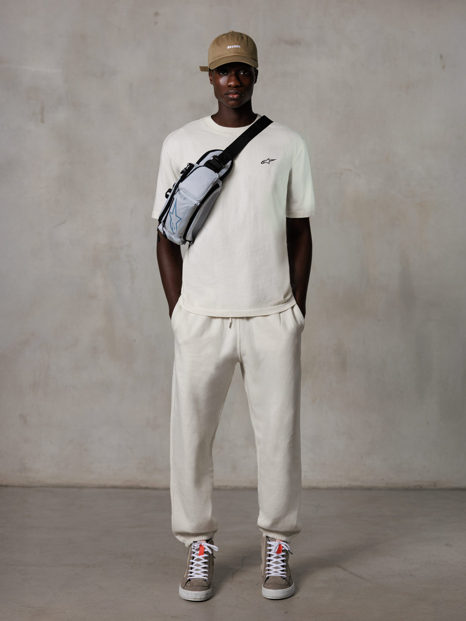 Alpinestars Essentials Ageless SS Tee, Short Sleeve T-Shirt, Off White, featuring a small black logo on the chest, worn with matching joggers, tan baseball cap, and a grey crossbody bag, casual lifestyle streetwear