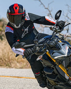 Motorcyclist leaning into a turn on a naked sport bike, wearing the Alpinestars T-SPS Air v2 Jacket in black with white and red fluo accents, paired with a black and red helmet and black gloves, performance-oriented textile gear for urban sport riding.