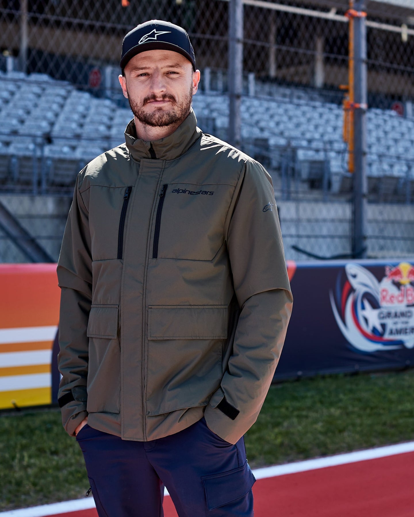 Jack Miller wearing a green Alpinestars jacket and cap standing on a race track with a blurred background
