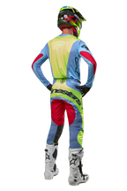Motocross rider from behind wearing 2024 Techstar Ocuri Pants and matching jersey, Light Blue Yellow Fluo and Red Berry, white and black boots, black helmet with neon accents, coordinated Alpinestars gear for off-road racing