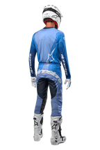 Motocross rider from behind wearing Alpinestars 2024 Techstar Pneuma Pants in Dark Navy and Light Blue, coordinated with a matching blue jersey, white helmet, white gloves, and white boots, featuring bold Alpinestars branding across the seat and sleeves.